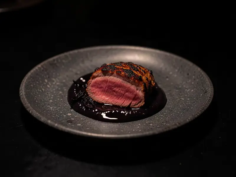 Sliced lamb loin, pink in the middle, on a dark plate with black garlic sauce