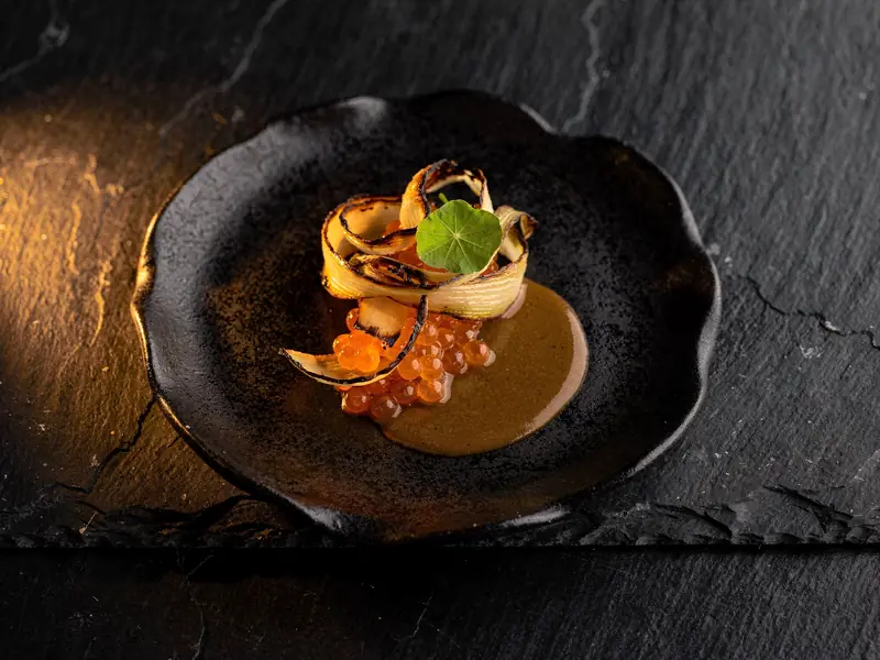 Charred leek ribbons with orange trout roe and brown butter on a black ceramic plate